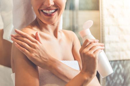 woman in bath towel applying body lotion on her shoulders and smiling while standing in bathroom